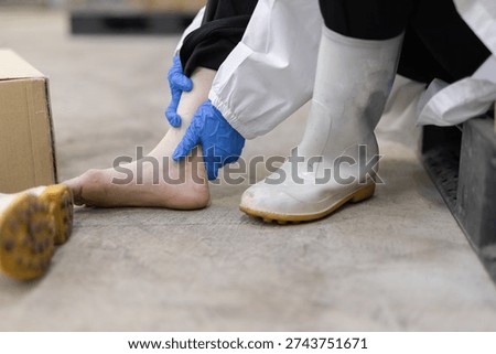 Factory worker experiencing ankle pain after removing safety boot in warehouse. Industrial workplace injury risk concept in manufacturing, logistics with cardboard boxes and pallet storage.