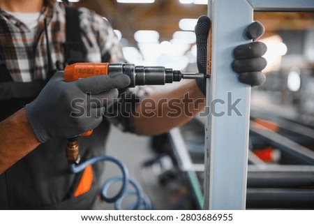 Factory worker in gloves using an air screwdriver, mounting an element of a white plastic window profile on an industrial assembly line
