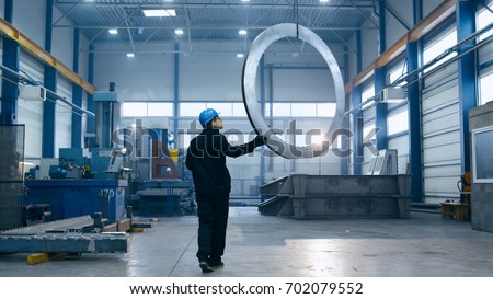Factory worker in a hard hat is moving a metal detail with a remote crane.