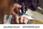 Factory worker making new shoes. Woman stitching details for white leather sneakers on industrial sewing machine, needle and hands holding material in closeup. Footwear manufacturing industry concept