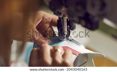 Factory worker making new shoes. Woman stitching details for white leather sneakers on industrial sewing machine, needle and hands holding material in closeup. Footwear manufacturing industry concept