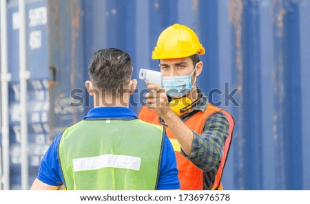 Factory worker man checking fever by digital thermometer for scan and protect from Coronavirus (COVID-19) at cargo containers - Healthcare Concept