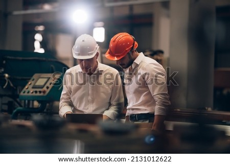 Factory worker man and engineer manager working together a metal sheet industrial worksite , checking machine process on laptop and wearing hard hat for safety