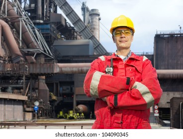 Factory Worker Posing In Front Of A Blast Furnace, Wearing Safety Gear, Including A Hard Hat, Goggles And Fire Retardant Coveralls