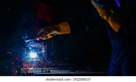 A factory worker wearing a green mechanic coveralls and safety helmet welding metalwork at night time in a factory while his colleague looking at a spark from welding.