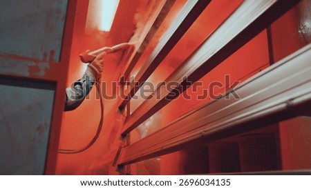Factory worker's gloved hand operates a spray gun, precisely applying vibrant orange powder coating to metal parts on a production rack in an industrial finishing line