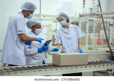 Factory workers review tablet data while standing beside conveyor and packaging machine. Concept shows digital monitoring, teamwork, and process control in manufacturing workflow.