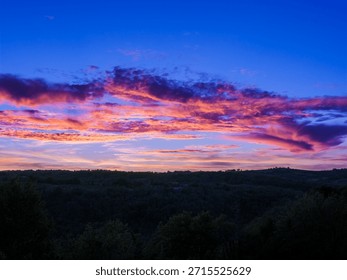Factual image of a dramatic sunset with vibrant orange and magenta clouds against a deep blue sky, viewed over a silhouetted forest line in Quercy, France.