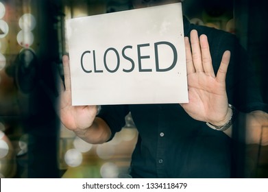 Failed Business Going Down Or Opening Times Concept. Man Putting Closed Sign In Window In Shop. Late At Night In City.