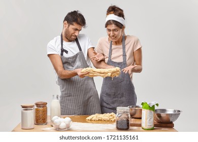 Failure And Cooking Concept. Disappointed Woman And Man Stretch Raw Sticky Dough, Have Some Problems With Making Homemade Pizza, Real Kitchen Disaster, Look With Disgusting Faces, Kitchenware On Table