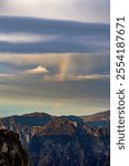 A faint rainbow arching above dramatic mountain peaks under cloudy sky at sunset . 