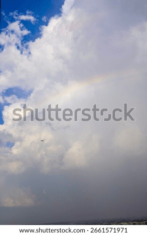 A faint rainbow arcs across a dramatic sky as storm clouds begin to clear over a distant cityscape. A small airplane flies beneath the clouds, adding a sense of scale and movement to the scene. 