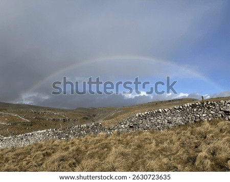 Faint rainbow on a half grey, half blue sky with fluffy clouds on the horizon. In the foreground, English countryside covered by winter’s golden grass. Rock walls throughout the landscape. 