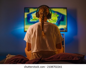 A fair-haired girl gamer sits in front of a large TV screen and plays a video game. Close-up. Shot from the back. Gaming, fun pastime, modern computer technologies, prizes, win.