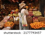 Fair-skinned young girl holds orange standing with her back to camera in fruit street shop. Brunette wears hat, blouse, pants and string bag. Concept shopping