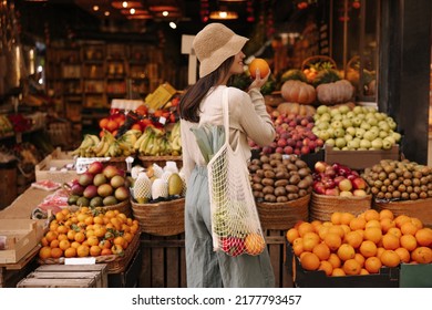 Fair-skinned young girl holds orange standing with her back to camera in fruit street shop. Brunette wears hat, blouse, pants and string bag. Concept shopping - Shutterstock ID 2177793457