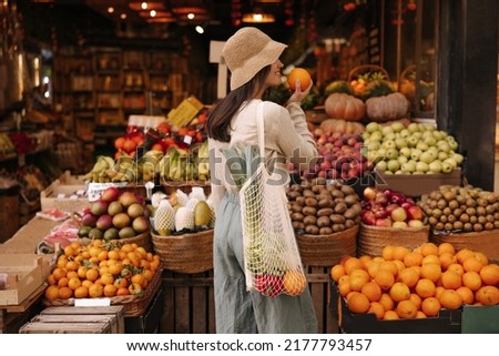 Fair-skinned young girl holds orange standing with her back to camera in fruit street shop. Brunette wears hat, blouse, pants and string bag. Concept shopping