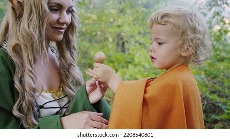 Fairy Elf Cosplay Woman Treats Little Baby Boy Hobbit With Candy In Green Forest. Halloween Concept, Fairytale Characters, Kids. High Quality Photo