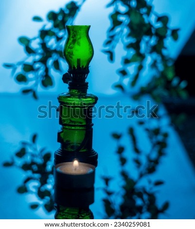 Fairy image of a transparent green glass lamp and a lit candle at its foot, backlit against a background of blurred branches and leaves reflecting on the dark shelf, creating a magical atmosphere