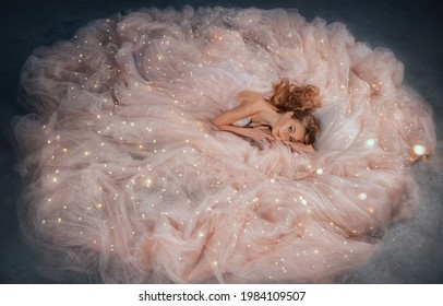 Fairy Queen Fashion Model In Luxurious Shining Pink Dress Posing In Studio. Princess Girl, Peach Outfit With Sequins Lies In Fabrics Of Outfit, Long Skirt Gown. Fantasy Woman Sleeping Beauty Woke Up