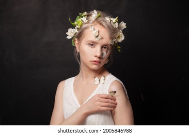 Fairy Tale Nymph Lady. Beautiful Young Blonde Female With White Spring Flowers In Her Hair And Butterfly On Her Healthy Skin On Black Background