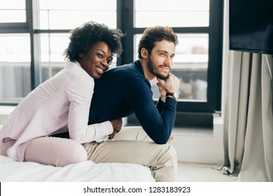 Faithful, Affectionate African Woman Is Sitting Behind And Hugging Tenderly Her Handsome Husband, They Watching Tv While Sit On The Bed In Spacious Room.