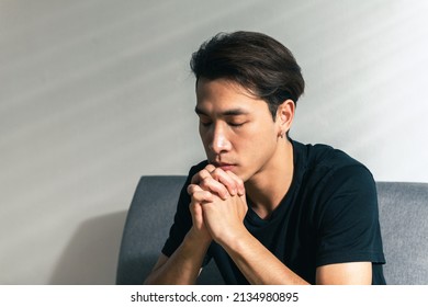 A faithful Asian young male hands in prayer gesture sitting alone on sofa at home office pray and hope for good luck, success, forgiveness. Power of religion, belief, god faith, worship concept ideas.