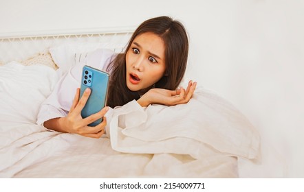 Fake news concept : Beautiful asian young woman wakes up from her bed looking at her smartphone, her eyes bulging and shocked by the spread of fake news that is so popular in the online community.