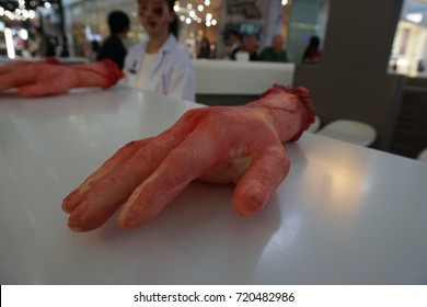 Fake Rubber Severed Hand With Terrible Blood, Halloween Prank Toy. Blurred Background Of Doctor Woman Halloween Makeup.