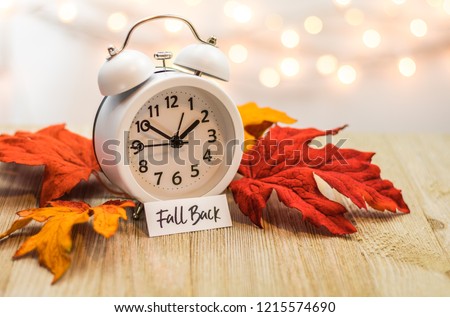 Fall Back Daylight Saving Time concept with white clock and autumn leaves, soft bokeh background on wooden board