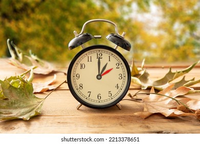 Fall Back one hour. Daylight Saving Time, Black alarm clock with time change on wooden table. Autumn trees and leaves background - Shutterstock ID 2213378651