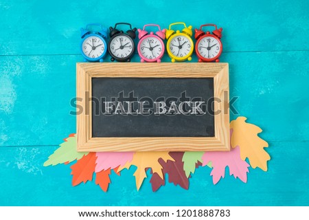 Fall back time change concept. Creative composition with alarm clock, autumn leaves and chalkboard. Top view from above