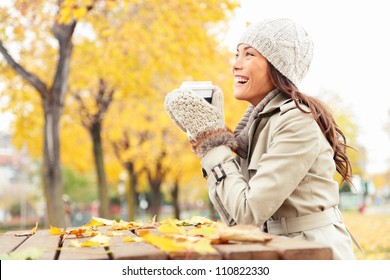 Fall concept - autumn woman drinking coffee on park bench under fall foliage. Beautiful young modern woman smiling happy and cheerful in trench coat.