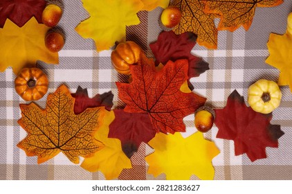 Fall leaves and tiny pumpkins rest on checkered fabric. Warm orange, red, and yellow hues dominate the scene. Decorative autumn leaves and gourds create cozy seasonal charm