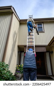 Fall Maintenance, Senior Man Climbing A Ladder With A Leaf Blower, Senior Woman Holding The Ladder Steady
