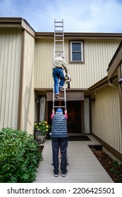 Fall Maintenance, Senior Man Climbing A Ladder With A Leaf Blower, Senior Woman Holding The Ladder Steady
