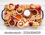 Fall pie theme dessert board. Overhead view table scene against a white wood background. Pumpkin, apple and pecan mini pies, with whipped cream, caramel and fruit. 