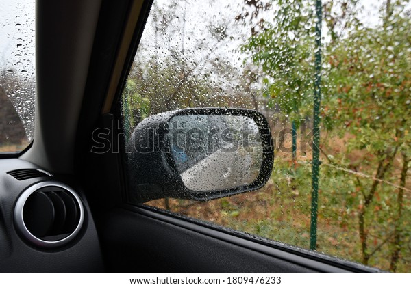 Fall rain shot inside car. Blurred\
background with wet side window of automobile in rainy weather.\
Defocus side mirror behind drizzled glass\
surface