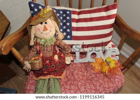 A fall scarecrow rests against an American Flag pillow.  In true farmhouse style, the cushioned rocking chair is filled with love.
