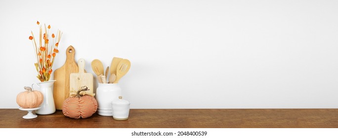 Fall theme kitchenware and utensils and decor on a wood shelf or counter. Banner against a white wall background with copy space. Autumn kitchen home cooking concept.
