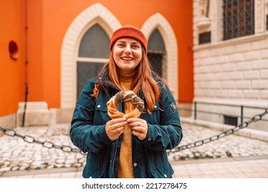 Fall Tourist Woman In A Bright Hat And Autumn Jacket Holding Baked Obwarzanek Traditional Polish Cuisine Snack Bagel On Old City Market Square In Krakow 