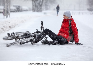 Fallen Down Caucasian Cyclist Sitting On Slippery Roadside, Riding Bicycle In City At Winter Season