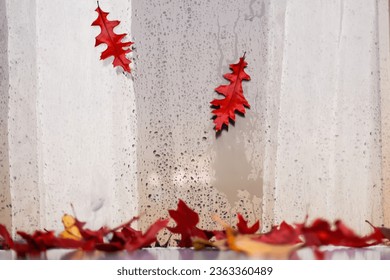 falling autumn dry red oak leaves against the background of a wet window with curtains, view from the street. Autumn concept of weather change