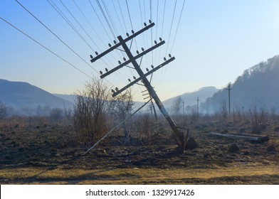 Falling electric pole in the field of the burned-down grass. The inclined pole is propped up by a branch. The grass in the field continues smokes. Mountains and  blue sky visible in the distance