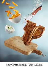 Falling T-bone steak meal with cutting board suspended in the air with a bowl of fried potato wedges, tzatziki dip, a sharp knife, salt and pepper over a graduated grey background with shadow below
