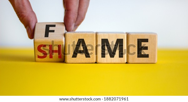 Fame or shame symbol. Male hand flips wooden\
cubes and changes the word \'shame\' to \'fame\' or vice versa.\
Beautiful yellow table, white background, copy space. Business and\
fame or shame concept.
