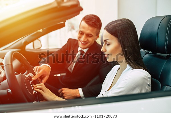 Familiarization with the design of a new car.\
Smiling salesman of dealership shows the car\'s functions to a young\
beautiful woman who sits behind the\
wheel.