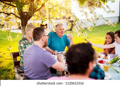 Familly Sitting Together At The Picnic Table And Eating Long Grilled Meat.