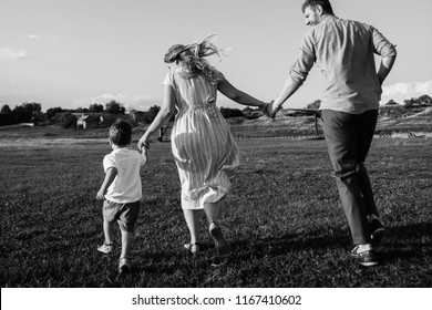 A Family Of 3 People, Father Mother And Son Run Across The Field Holding Each Other Hands. Photo From Behind Black And White Photo. Happy Young Family Spending Time Together Outside In  Nature