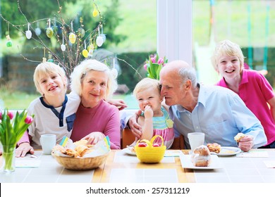 Family Of 5: Grandparents And Grandchildren, Teenage Boys And Toddler Girl Eating Eggs And Pastry Enjoying Family Breakfast On Easter Day Sitting Together In Sunny Kitchen With Big Garden View Window